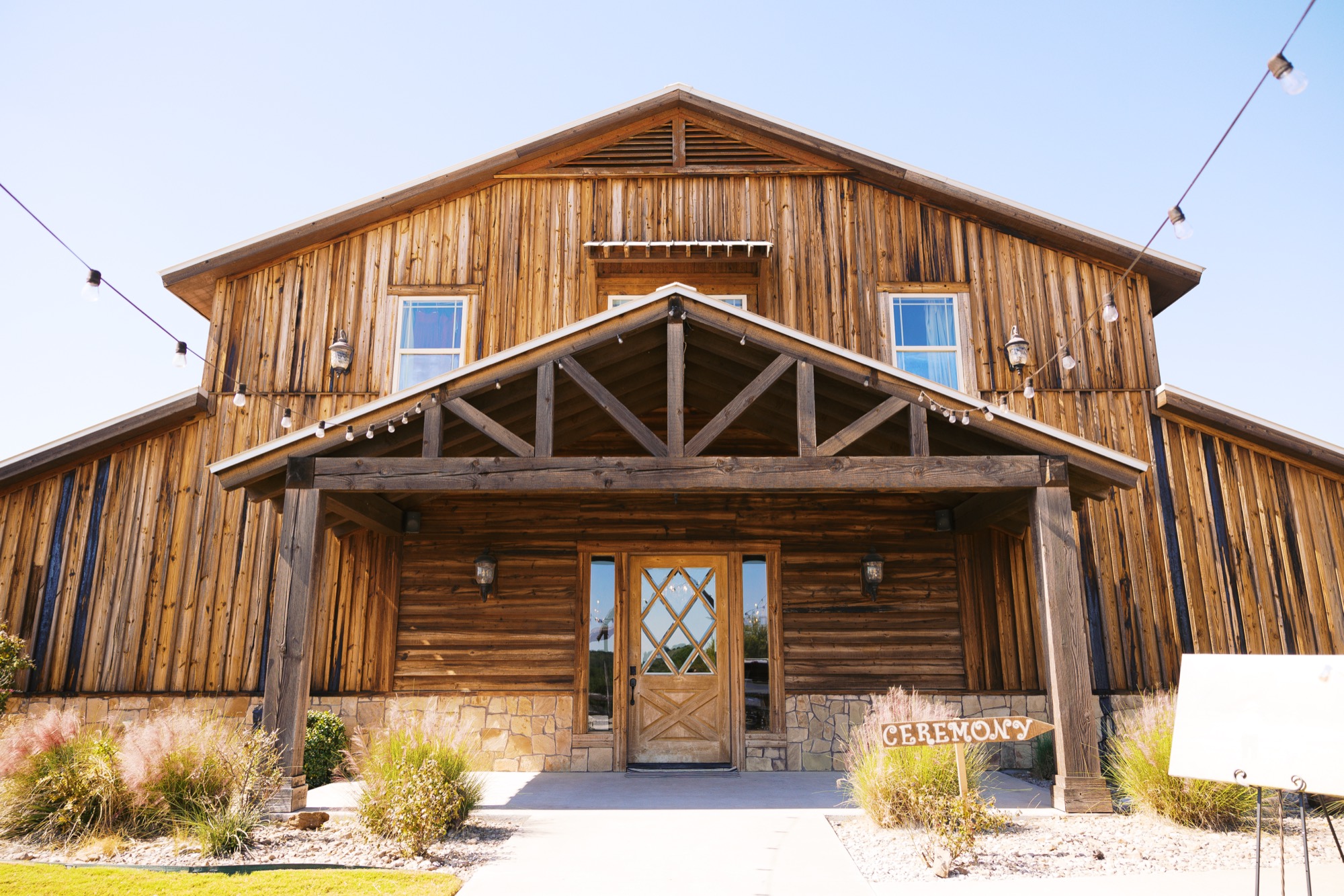 The front facade of the timber reception barn at Diamond H3 Ranch, with its covered entrance, glass doors, string lights, and stone base