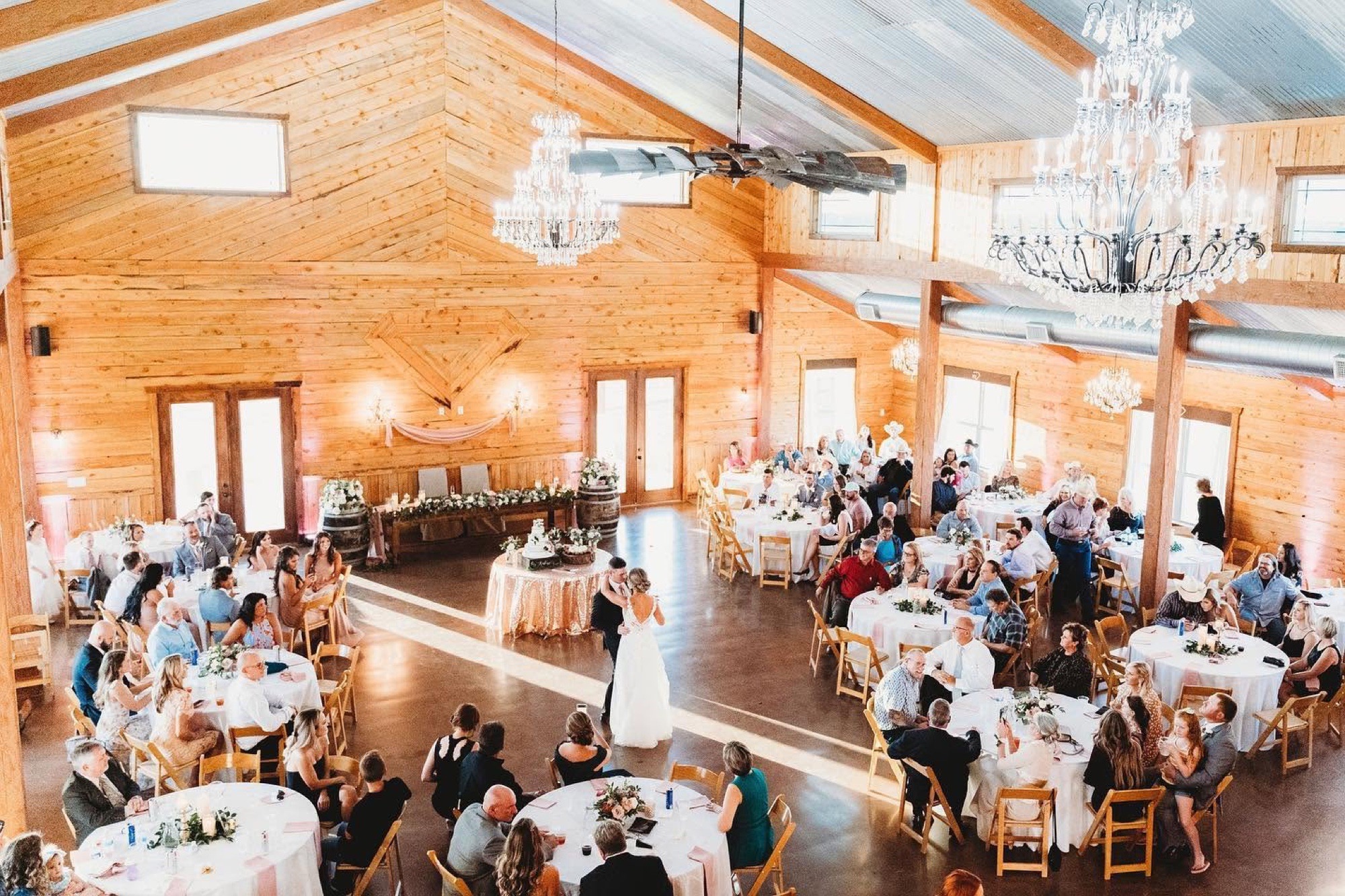 A wedding reception inside the cedar barn at Diamond H3 Ranch — round tables, two crystal chandeliers, and the couple's first dance