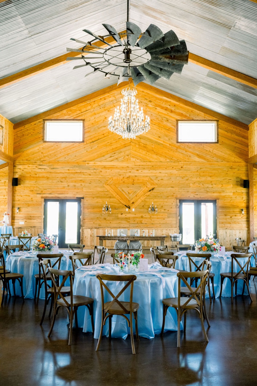 The reception barn interior at Diamond H3 Ranch, round tables with light-blue linens beneath a windmill chandelier and cathedral tin ceiling