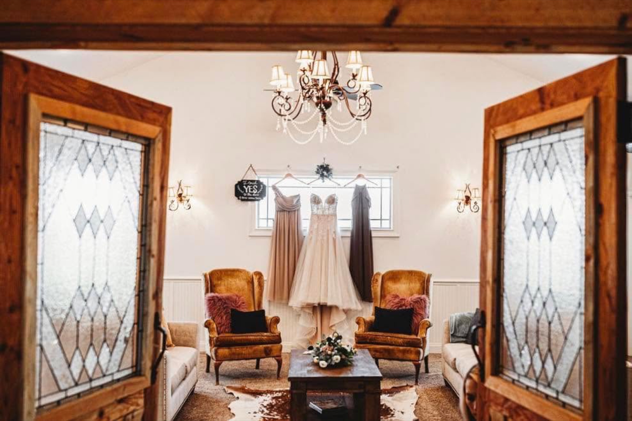 The bridal suite lounge at Diamond H3 Ranch, framed by leaded-glass doors, with a chandelier, velvet chairs, and the wedding dress hanging